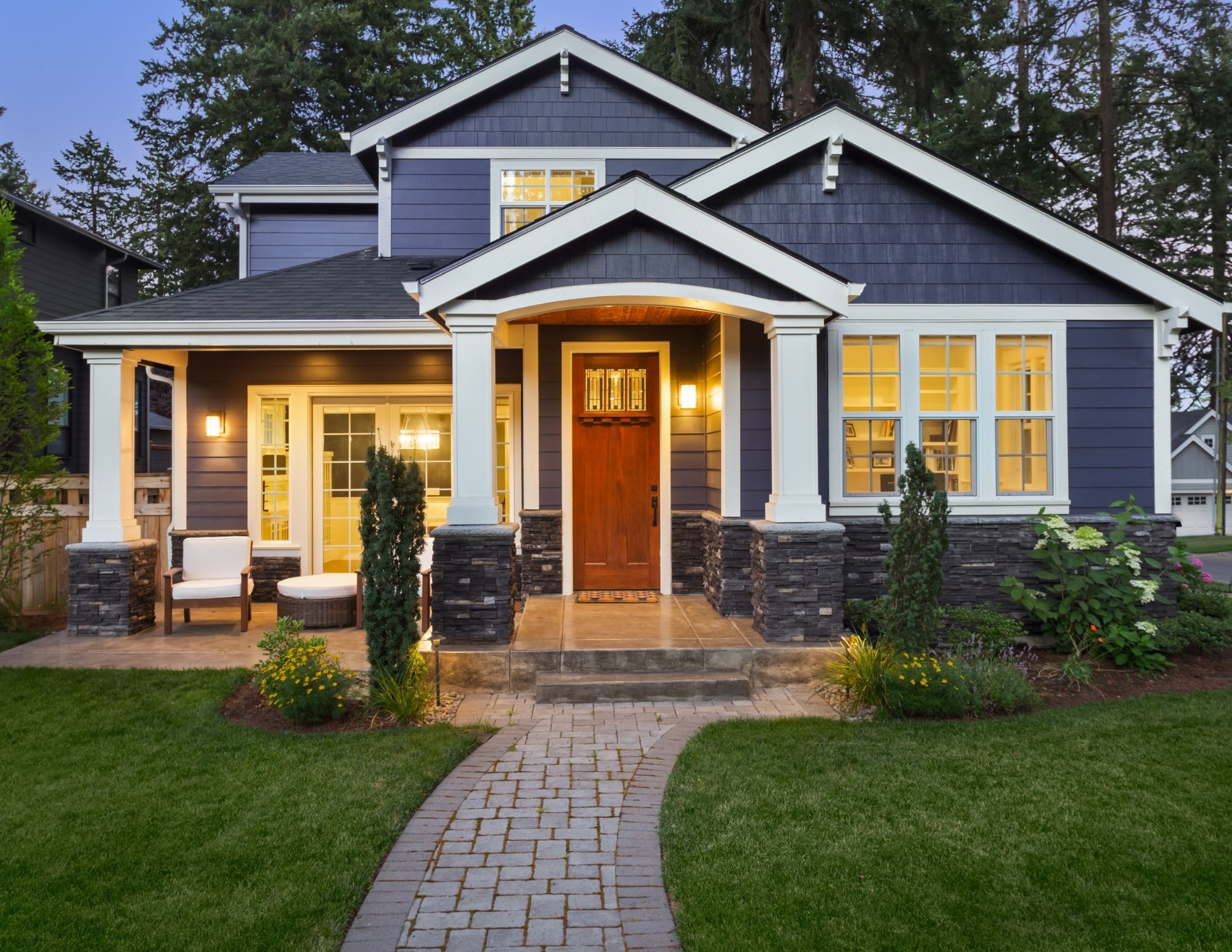 Modern two-story home with dark blue siding, white columns, and warm lighting illuminating the red front door and large windows at dusk