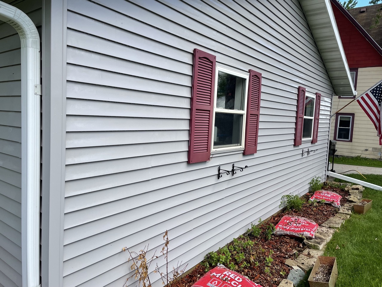 White vinyl siding house with burgundy shutters and windows, featuring landscaping beds with red mulch and protective covers along the foundation