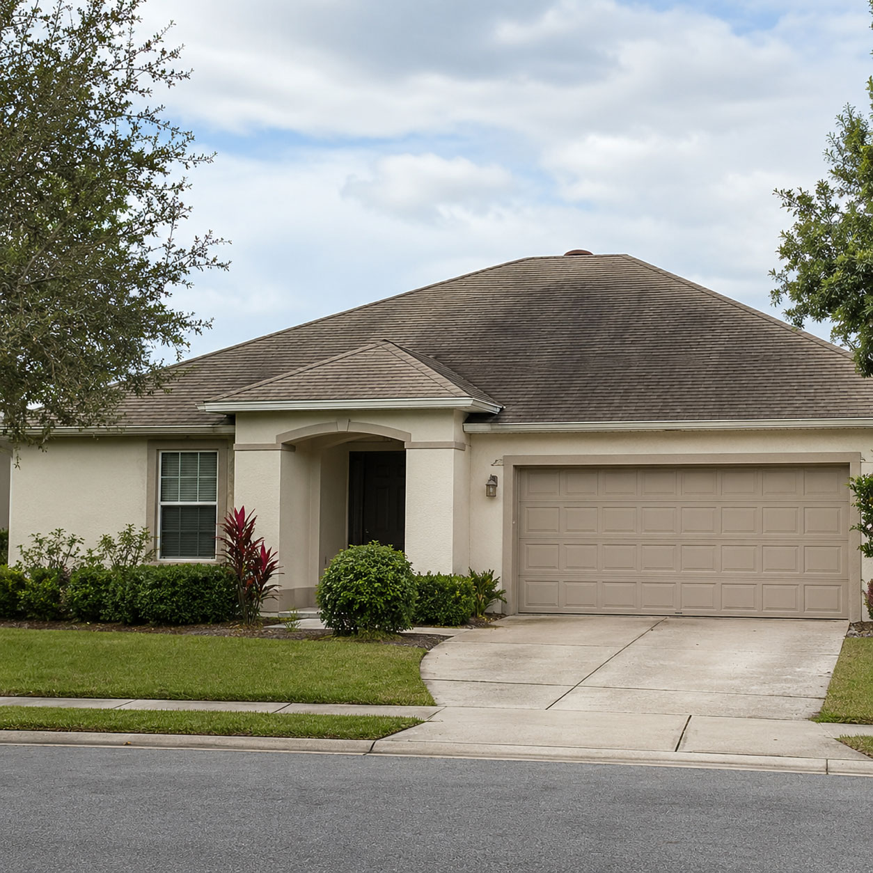 Single-story beige house with uneven gray roof coloring, tan garage door, manicured lawn, and red tropical plants near entrance in Fond du Lac, Wisconsin