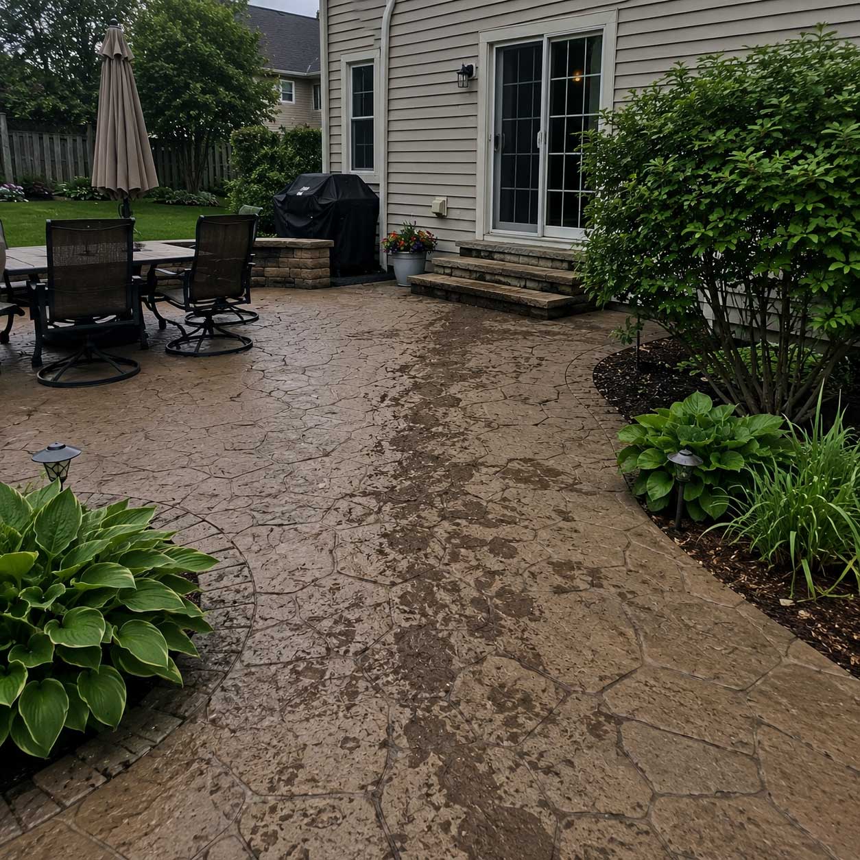 Dirty residential patio before pressure washing, with mud stains on stamped concrete surface surrounded by landscaping and house