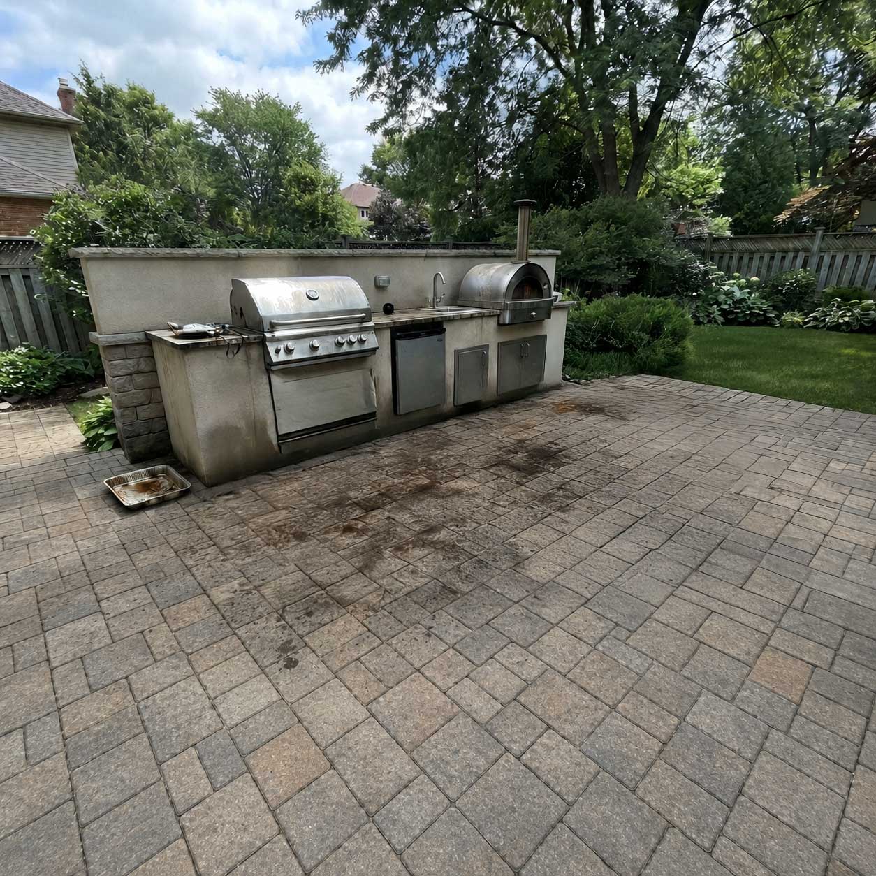 Outdoor patio with dark grease stains on concrete pavers next to a built-in stainless steel grill and pizza oven in a residential backyard