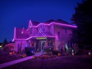 A Victorian house in Fond du Lac, Wisconsin brilliantly decorated with bright magenta LED lights outlining the roofline and forming a ribbon bow design, surrounded by autumn pumpkins and corn stalks