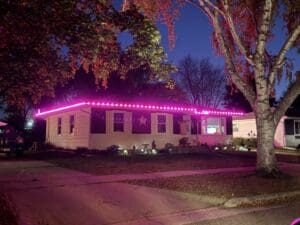 A mobile home at night illuminated by bright pink neon lights along the roofline, with autumn foliage overhead and a star decoration visible on the front panel