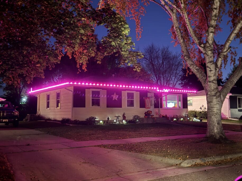 A mobile home at night illuminated by bright pink neon lights along the roofline, with autumn foliage overhead and a star decoration visible on the front panel