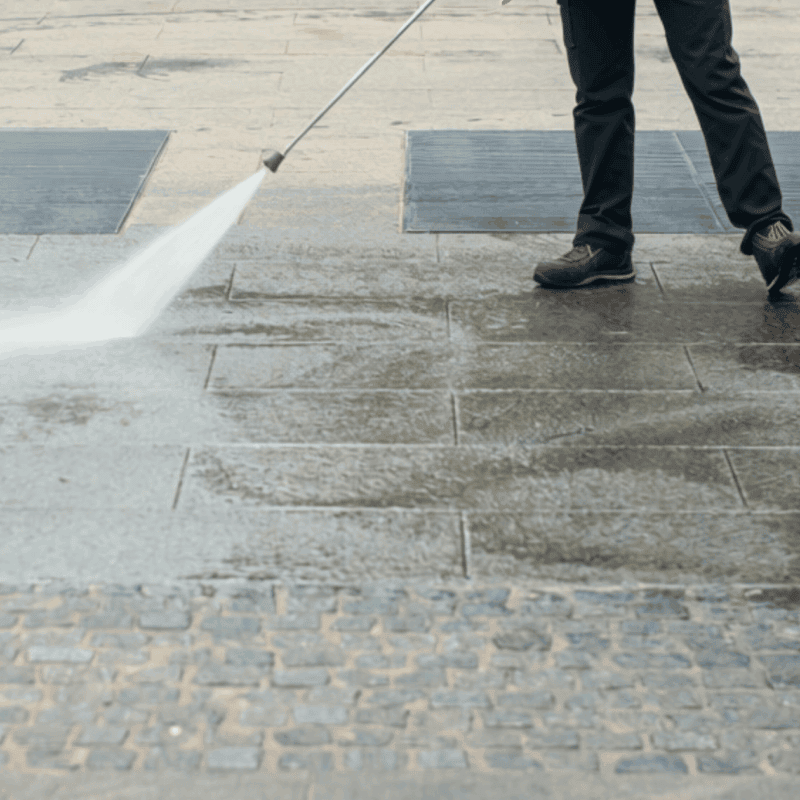Person pressure washing concrete sidewalk and patio area with high-pressure water stream, removing dirt and stains from residential or commercial exterior surface