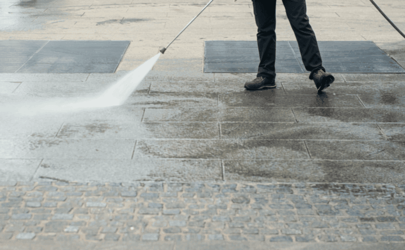 Person pressure washing concrete sidewalk and patio area with high-pressure water stream, removing dirt and stains from residential or commercial exterior surface