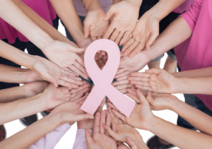 Multiple hands in pink clothing circle a pink breast cancer awareness ribbon with a heart cutout