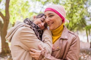 Woman wearing pink headwrap and mustard turtleneck holds smiling child outdoors under tree canopy in autumn setting